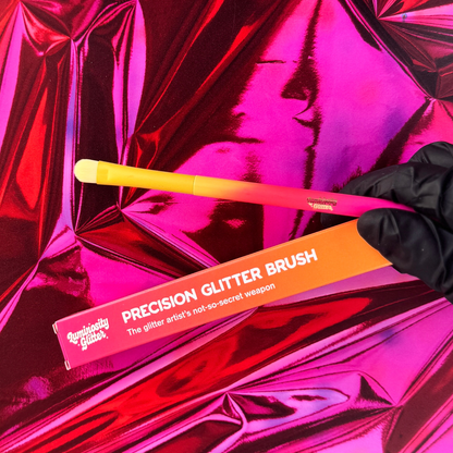 Precision glitter brush with orange handle and packaging on pink reflective background