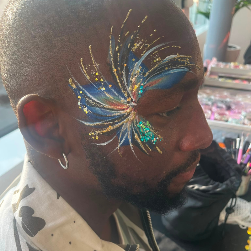 A close-up side profile of a man with elaborate face paint featuring a blue, gold, and white burst design on his forehead and cheek, with a small teal glitter accent.