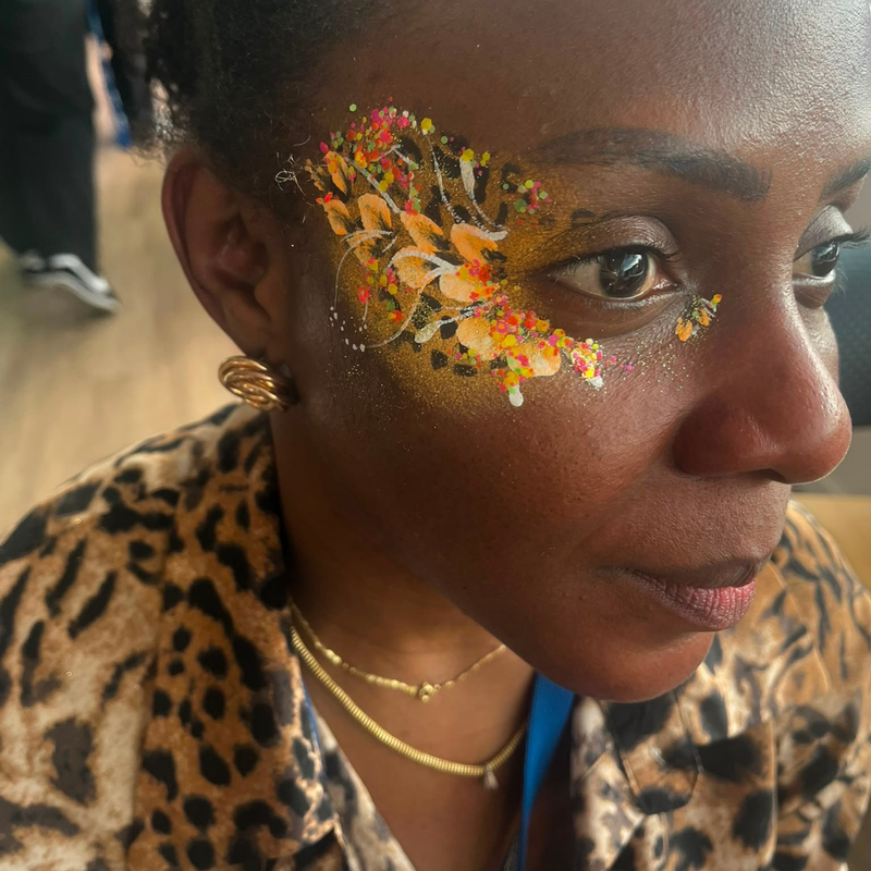 A close-up of a woman wearing a leopard print jacket and gold jewelry, featuring a colorful face paint design on her temple with autumn leaves and splatters.