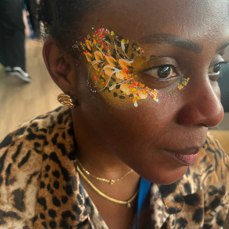A close-up of a woman wearing a leopard print jacket and gold jewelry, featuring a colorful face paint design on her temple with autumn leaves and splatters.