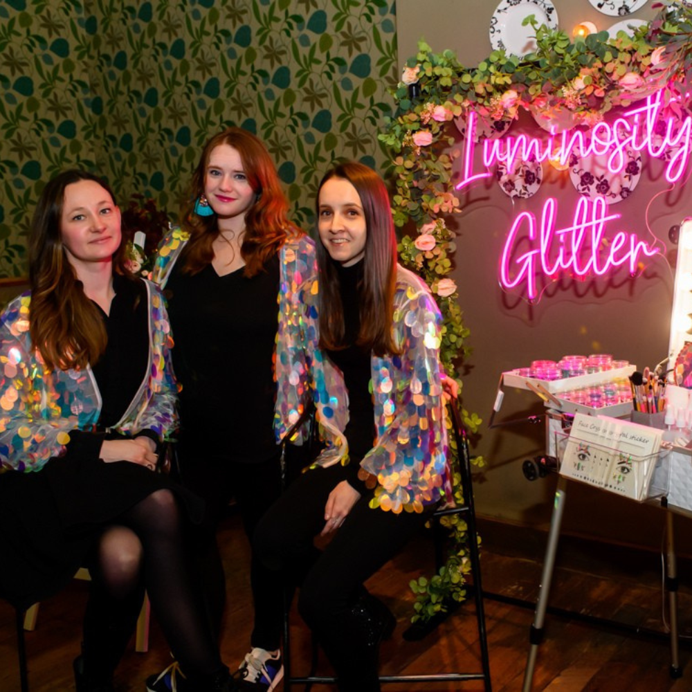 Three women wearing shiny multicolor sequined jackets pose in front of a pink neon sign that reads 'Luminosity Glitter' in a decorated room with floral arrangements and patterned wallpaper.