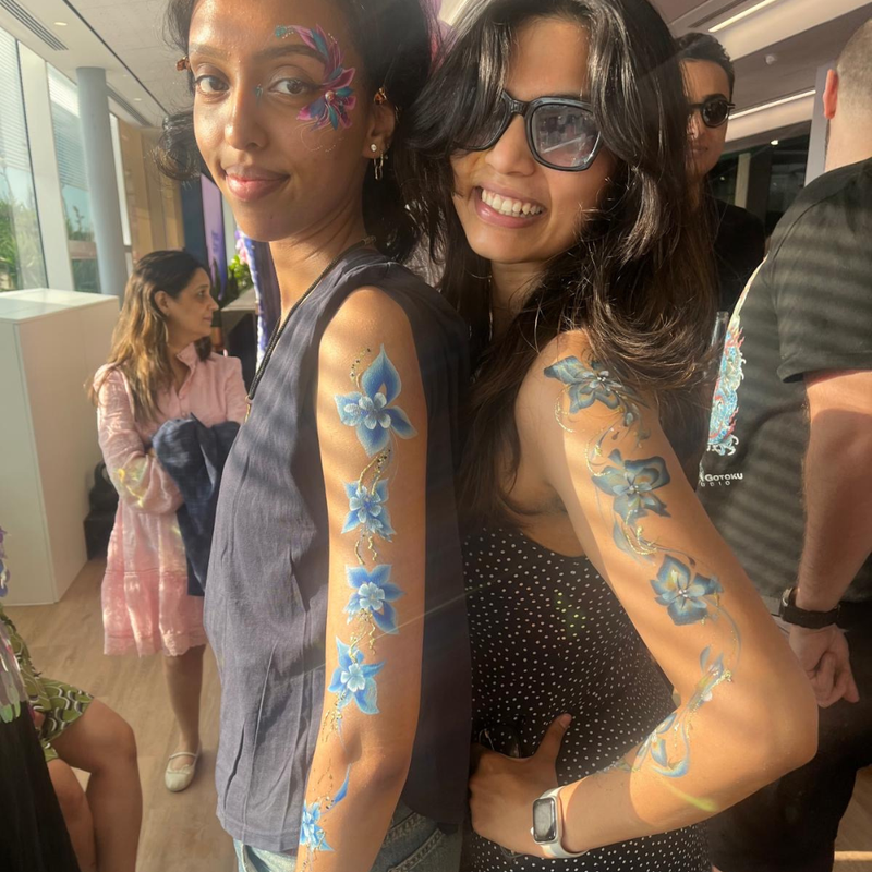 Two women posing together indoors, showcasing temporary body paint designs on their arms. The woman on the left has blue floral and crescent moon designs painted on her arm, while the woman on the right has matching blue floral designs on her upper arm and forearm.