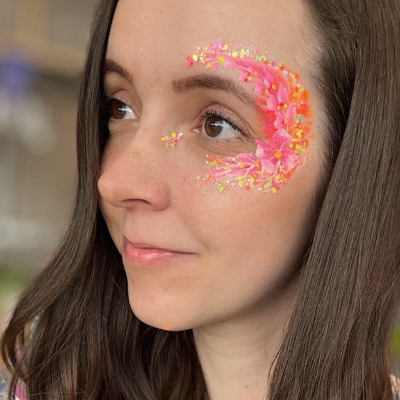A close-up of a woman's face showing a decorative pink and orange floral face paint design with yellow glitter accents on her left cheek.