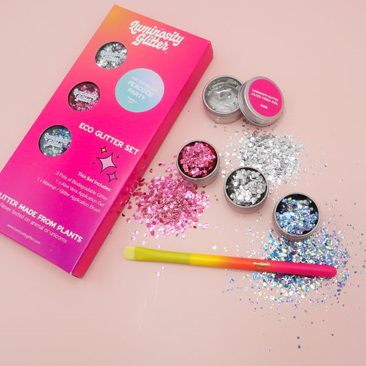 The image shows a collection of biodegradable cosmetic glitter in various pots with a brush and application gel, displayed alongside their packaging box, all set against a pink background.