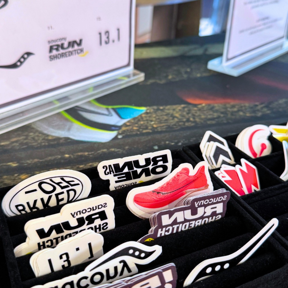 A close-up of a display of Saucony temporary tattoos in a black tray, featuring various designs including a red sneaker, white text stickers, and black-and-white logo stickers.